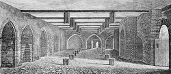 A monochrome illustration of a stone and brick-walled room.  An open doorway is to the right.  The left wall contains equally spaced arches.  The right wall is dominated by a large brick arch.  Three arches form the third wall, in the distance.  The floor and ceiling is interrupted by regularly spaced hexagonal wooden posts.  The ceiling is spaced by wooden beams.