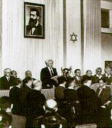 A man reads a document to a small audience assembled before him. Behind him are two elongated flags bearing the Star of David and portrait of a bearded man in his forties.