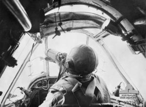 Pilot in a small cockpit. He is wearing a parachute and leather helmet. This is a still from movie footage shot by Damien Parer.