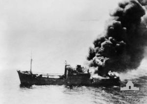 A ship burning from the stern settles in the water.