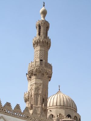 An ornate carved stone minaret, with a carved stone railing around three balconies, the first below its center, the second two thirds the way up, and the third near its top. The tip of the minaret is a large bulb-shaped stone decoration with a small bulb-shaped metal finial. Behind the minaret most of a dome is visible.