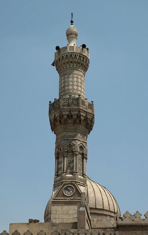 An ornate carved stone minaret, with a carved stone railing around balconies at its center and near its top. The tip of the minaret is a large bulb-shaped stone decoration with a small bulb-shaped metal finial. Behind the minaret part of the top of a dome is visible.