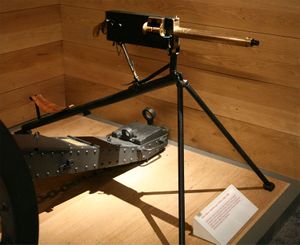 Brass barrelled gun with black coloured rear parts. On a tripod on a wooden plinth with a wood wall behind.