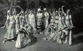 A group of female dancers in flowing robes strike exaggerated poses around a central group of two men and a woman.