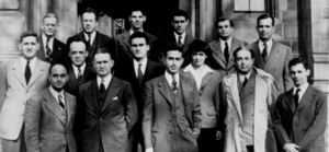 A group photo of 14 men in suits and 1 woman in three rows facing the vewier.