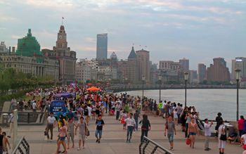 A wide curving walkway along a body of water to the right, crowded with many people and some large orange umbrellas underneath a sky colored by sunset. To the left of the walkway are light brownish-grey stone buildings in various late-19th and early-20th-century architectural styles, with taller buildings in the distance.