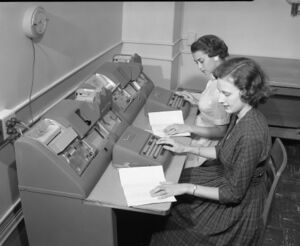 Black and white photo of two women sitting at desks with a punch machines