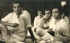 Photograph of Satyajit Ray seated with Ravi Shankar with several others in the background