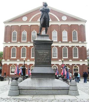 A statue on a pedestal of a man standing with his arms crossed. An inscription on the pedestal reads, "Samuel Adams, 1722–1803. A Patriot. He organized the Revolution and signed the Declaration of Independence." Behind the statue is a three story brick building with many windows.