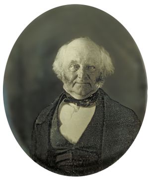 Half-length photographic portrait of an elderly, balding man dressed in a dark coat, vest and cravat